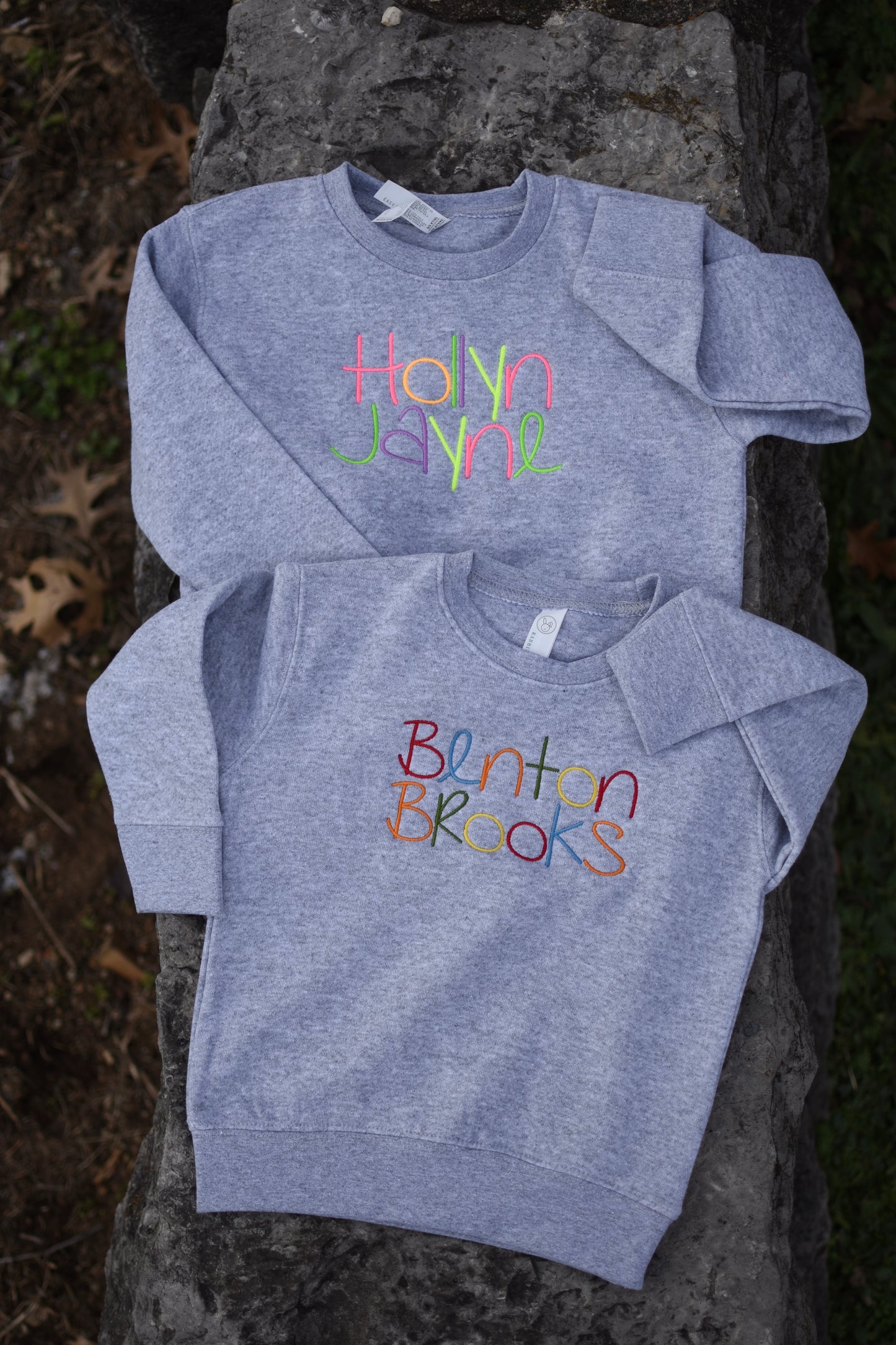 Two gray sweatshirts with colorful text on the front, one with the name 'Hayden' and the other with the name 'Benton'. Both are placed on a stone surface outdoors.