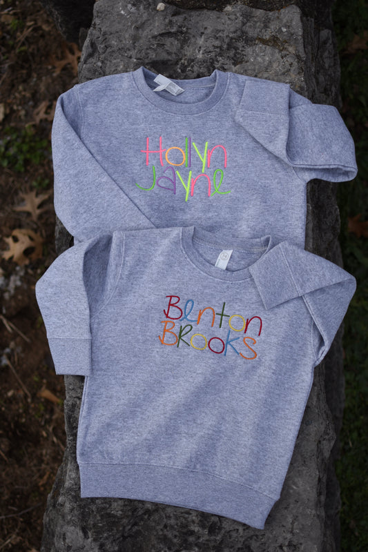Two gray sweatshirts with colorful text on the front, one with the name 'Hayden' and the other with the name 'Benton'. Both are placed on a stone surface outdoors.